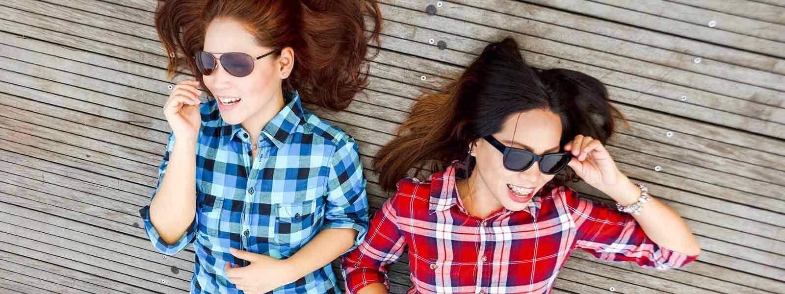 Two women in plaid shirts and sunglasses lying on wooden floor, laughing.