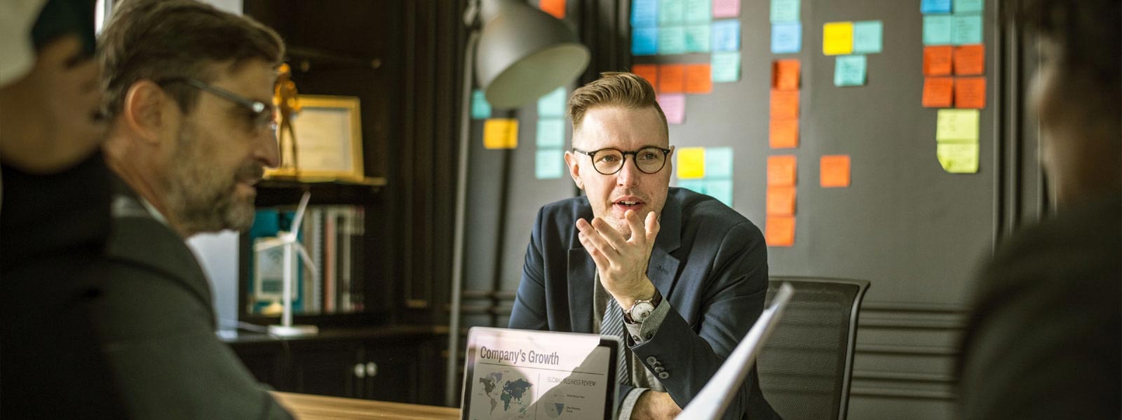 A man in glasses gestures while speaking in an office setting.